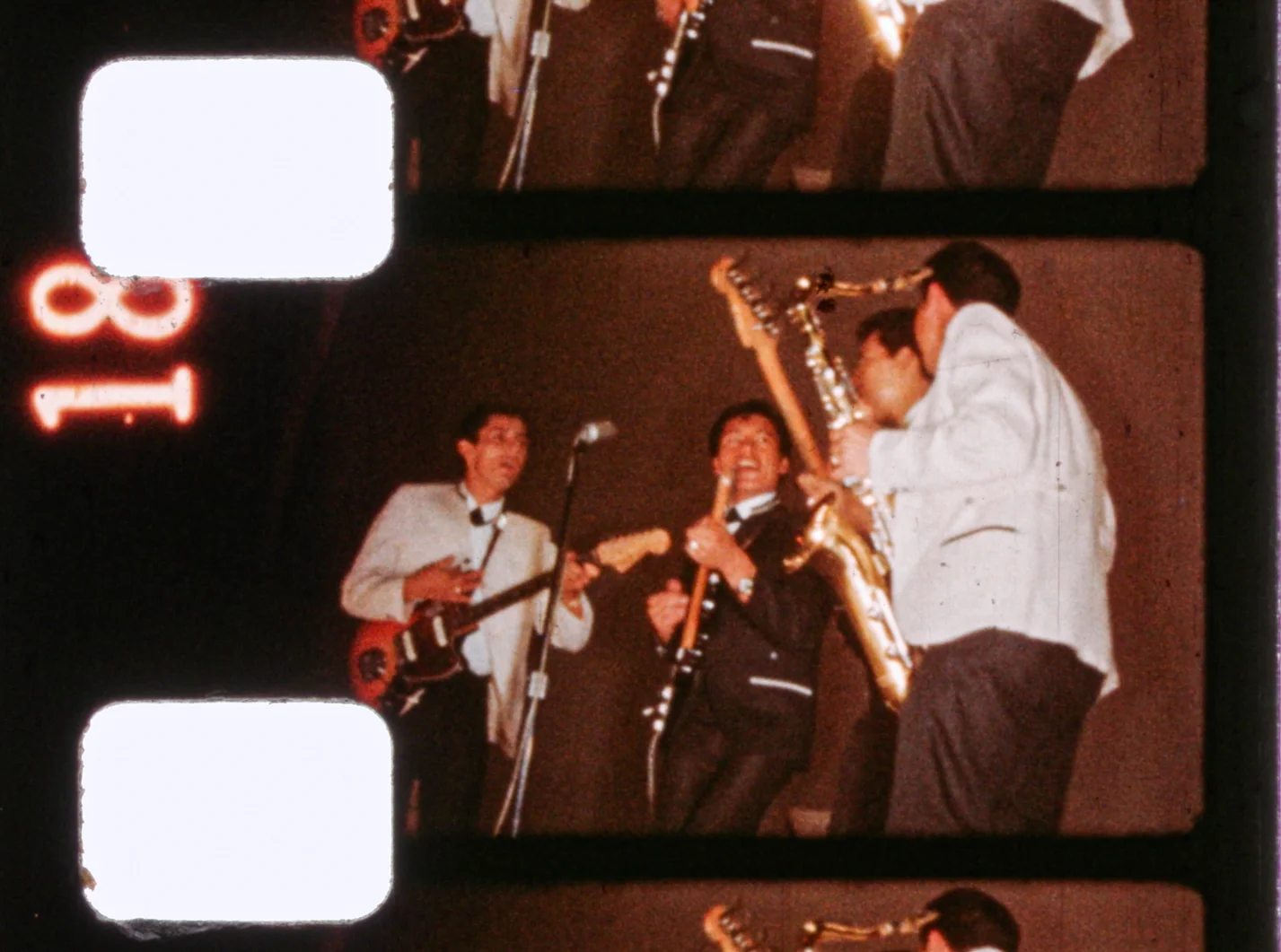 Fotograma de 8mm con el número de borde 18. Dos guitarristas, uno con saco claro y otro oscuro, cantan frente a un micrófono mientras un saxofonista toca a la derecha.