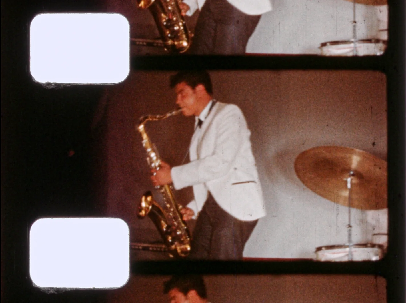 Fotograma de 8mm. Un joven con saco blanco toca el saxofón tenor de perfil, junto a un platillo de batería.
